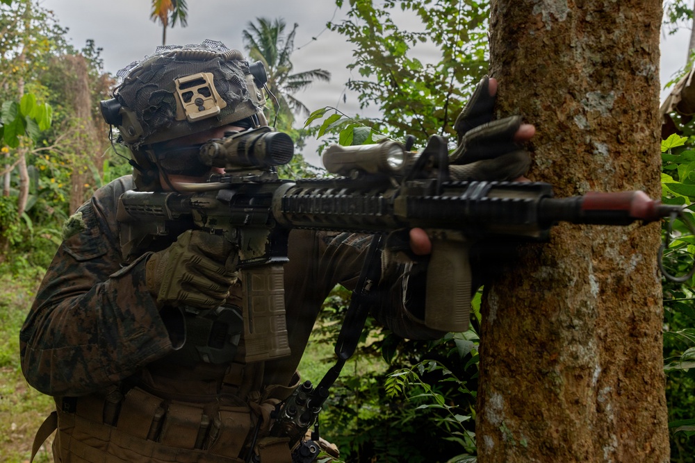 A soldier in full combat gear is taking a firing position behind a tree in a tropical environment. The individual is aiming down the scope of an assault rifle while using the tree as cover. The setting appears to be a jungle or tropical region based on the palm trees and dense vegetation visible in the background.
