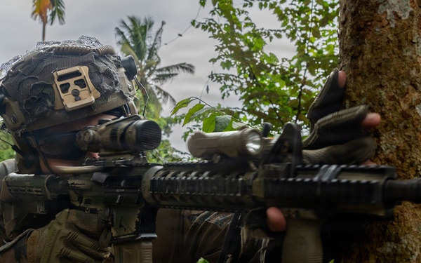 A soldier in full combat gear is taking a firing position behind a tree in a tropical environment. The individual is aiming down the scope of an assault rifle while using the tree as cover. The setting appears to be a jungle or tropical region based on the palm trees and dense vegetation visible in the background.