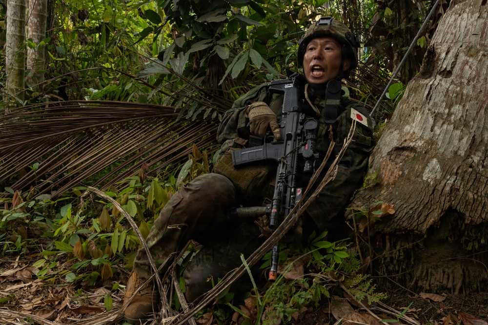 A soldier wearing Japanese Ground Self-Defense Force (JGSDF) woodland camouflage uniform is positioned against a large tree trunk in a dense tropical jungle environment. The soldier is holding an assault rifle and appears to be shouting or issuing commands, suggesting an active training exercise or tactical operation. A Japanese national flag patch is clearly visible on the left shoulder of the uniform.