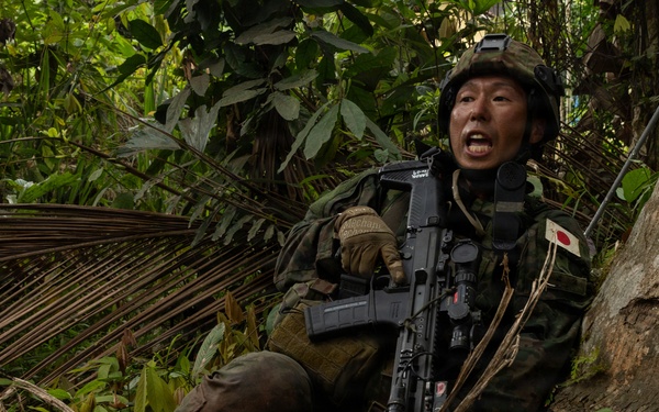 A soldier wearing Japanese Ground Self-Defense Force (JGSDF) woodland camouflage uniform is positioned against a large tree trunk in a dense tropical jungle environment. The soldier is holding an assault rifle and appears to be shouting or issuing commands, suggesting an active training exercise or tactical operation. A Japanese national flag patch is clearly visible on the left shoulder of the uniform.