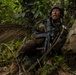 A soldier wearing Japanese Ground Self-Defense Force (JGSDF) woodland camouflage uniform is positioned against a large tree trunk in a dense tropical jungle environment. The soldier is holding an assault rifle and appears to be shouting or issuing commands, suggesting an active training exercise or tactical operation. A Japanese national flag patch is clearly visible on the left shoulder of the uniform.