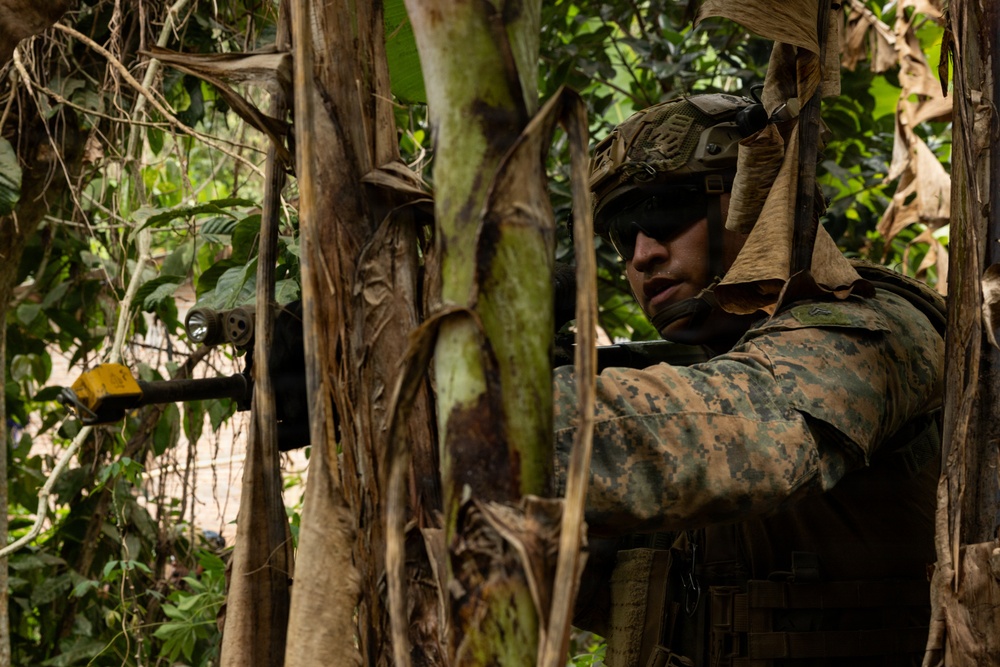 A U.S. Marine in full combat gear is positioned in dense jungle vegetation, aiming a rifle with a mounted flashlight and yellow training/blank-firing adapter attached to the muzzle. The Marine is wearing MARPAT (Marine Pattern) digital camouflage uniform, a tactical helmet with accessories, and body armor. The setting appears to be a jungle training exercise based on the lush tropical foliage and the presence of training equipment on the weapon.