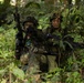 Two soldiers in woodland camouflage uniforms are positioned in dense jungle vegetation, appearing to be conducting a military training exercise or patrol. Both are armed and equipped with tactical gear including helmets, body armor, and communication equipment. Their posture and positioning suggest they are taking cover or conducting a reconnaissance halt.