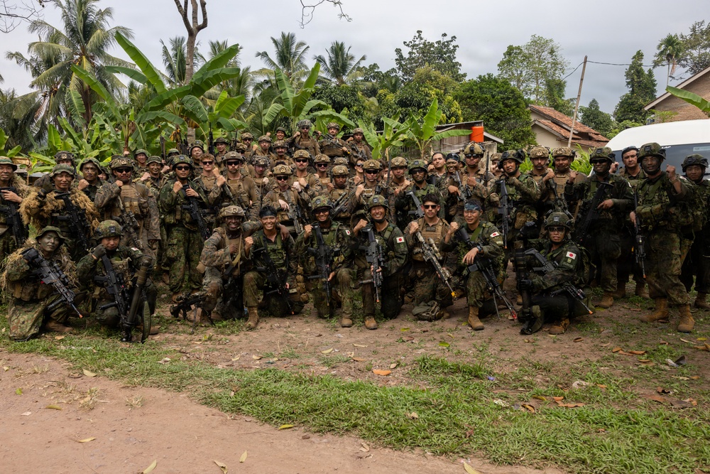 A large group of military personnel from at least two nations pose together for a group photograph in a tropical outdoor setting. Several soldiers are visibly wearing Japanese flag patches on their uniforms, suggesting this is a joint military exercise or training operation. The group is armed and in full combat gear, smiling and appearing in good spirits.