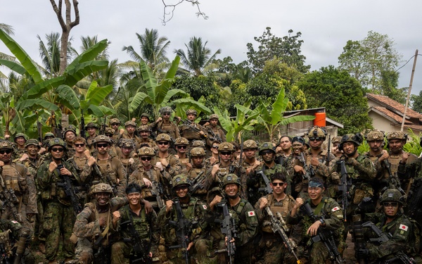 A large group of military personnel from at least two nations pose together for a group photograph in a tropical outdoor setting. Several soldiers are visibly wearing Japanese flag patches on their uniforms, suggesting this is a joint military exercise or training operation. The group is armed and in full combat gear, smiling and appearing in good spirits.