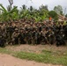 A large group of military personnel from at least two nations pose together for a group photograph in a tropical outdoor setting. Several soldiers are visibly wearing Japanese flag patches on their uniforms, suggesting this is a joint military exercise or training operation. The group is armed and in full combat gear, smiling and appearing in good spirits.
