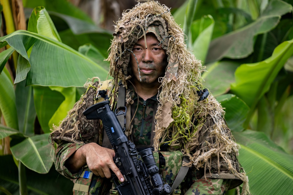 A soldier or military operative is photographed in a jungle or tropical environment wearing a ghillie suit over camouflage uniform with face paint applied. The subject is holding a rifle with an optical scope and is posed directly facing the camera. The setting features large tropical leaves, consistent with a Southeast Asian or Pacific Island environment.
