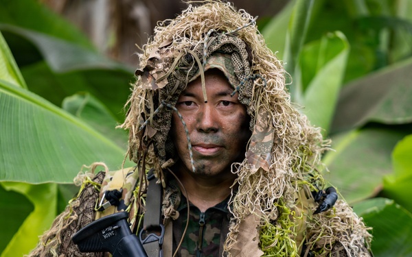 A soldier or military operative is photographed in a jungle or tropical environment wearing a ghillie suit over camouflage uniform with face paint applied. The subject is holding a rifle with an optical scope and is posed directly facing the camera. The setting features large tropical leaves, consistent with a Southeast Asian or Pacific Island environment.