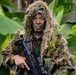 A soldier or military operative is photographed in a jungle or tropical environment wearing a ghillie suit over camouflage uniform with face paint applied. The subject is holding a rifle with an optical scope and is posed directly facing the camera. The setting features large tropical leaves, consistent with a Southeast Asian or Pacific Island environment.