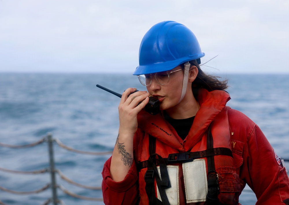 A person wearing a blue hard hat, safety glasses, and a red life vest/survival suit is speaking into a handheld radio (walkie-talkie) while standing on the deck of a vessel at sea. The background shows open ocean water and a rope railing along the ship's edge. The individual also has a visible tattoo on their forearm.