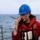 A person wearing a blue hard hat, safety glasses, and a red life vest/survival suit is speaking into a handheld radio (walkie-talkie) while standing on the deck of a vessel at sea. The background shows open ocean water and a rope railing along the ship's edge. The individual also has a visible tattoo on their forearm.