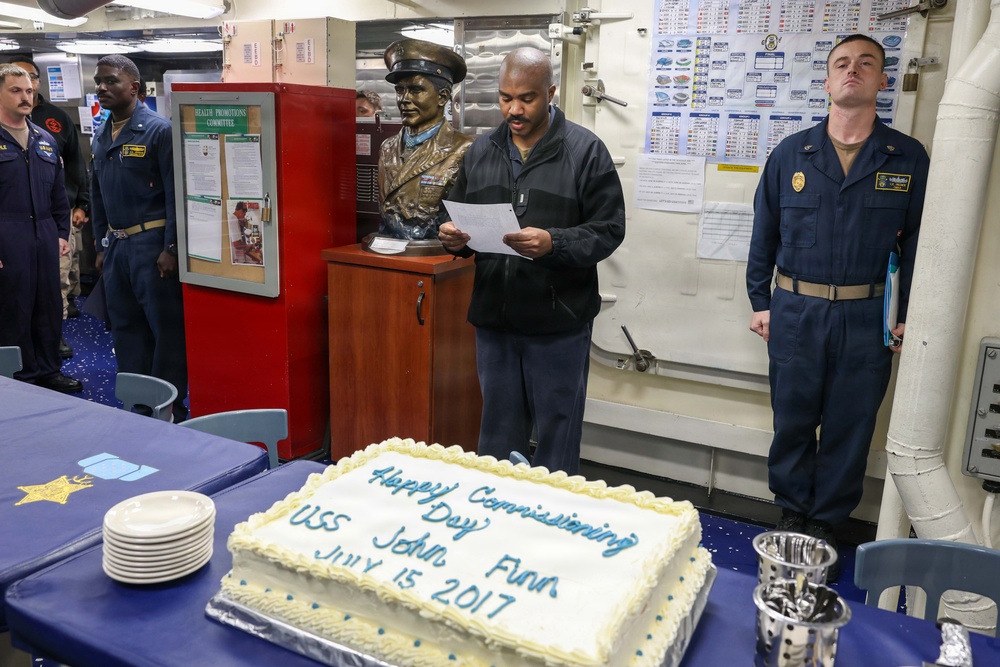 A commissioning ceremony is taking place aboard a U.S. Navy vessel, marked by a large decorated cake reading 'Happy Commissioning Day USS John Finn July 15 2017'. A man in civilian attire is reading from a paper near a bronze bust, while uniformed Navy personnel stand at attention. The setting appears to be an interior compartment of a naval ship.