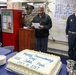A commissioning ceremony is taking place aboard a U.S. Navy vessel, marked by a large decorated cake reading 'Happy Commissioning Day USS John Finn July 15 2017'. A man in civilian attire is reading from a paper near a bronze bust, while uniformed Navy personnel stand at attention. The setting appears to be an interior compartment of a naval ship.