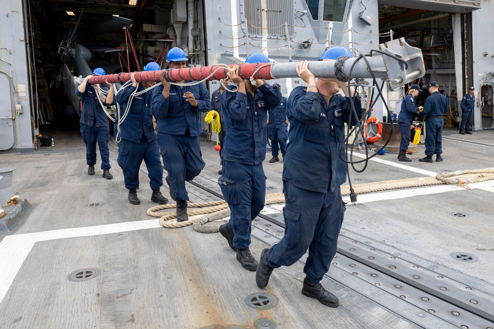 A group of U.S. Navy sailors in navy blue coveralls and blue hard hats are carrying a large red-and-silver cylindrical object, likely a torpedo or similar ordnance/equipment, across the deck of a naval vessel. Additional personnel are visible in the background performing various tasks near an open hangar bay, which contains what appears to be a helicopter. The scene depicts routine shipboard operations or weapons/equipment handling on what appears to be a large surface combatant or amphibious ship.