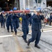 A group of U.S. Navy sailors in navy blue coveralls and blue hard hats are carrying a large red-and-silver cylindrical object, likely a torpedo or similar ordnance/equipment, across the deck of a naval vessel. Additional personnel are visible in the background performing various tasks near an open hangar bay, which contains what appears to be a helicopter. The scene depicts routine shipboard operations or weapons/equipment handling on what appears to be a large surface combatant or amphibious ship.