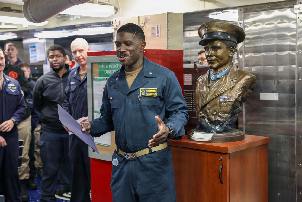 A U.S. Navy officer, identified by his patch as a Commanding Officer, addresses a group of personnel aboard what appears to be a naval vessel. He is standing next to a bronze bust of a military figure displayed on a wooden pedestal, likely during a ceremony or commemorative event. Several sailors and personnel are gathered in the background near a 'Health Promotion Committee' bulletin board.