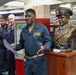 A U.S. Navy officer, identified by his patch as a Commanding Officer, addresses a group of personnel aboard what appears to be a naval vessel. He is standing next to a bronze bust of a military figure displayed on a wooden pedestal, likely during a ceremony or commemorative event. Several sailors and personnel are gathered in the background near a 'Health Promotion Committee' bulletin board.