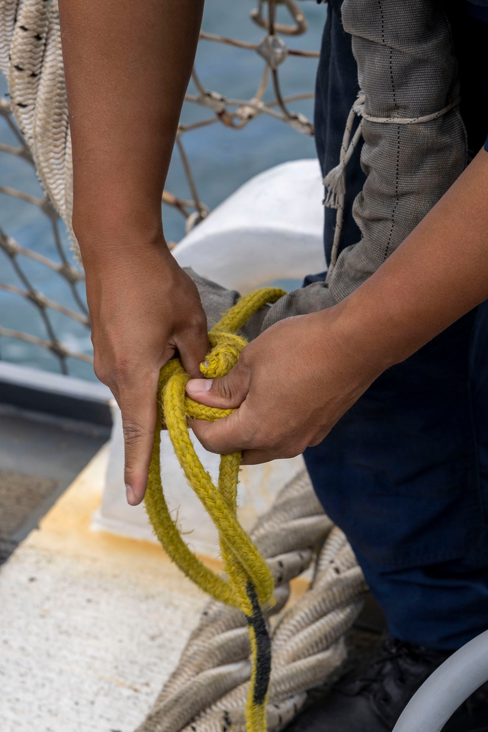 A person in navy blue pants and a gray vest is working with a yellow braided rope on what appears to be the deck of a vessel. They are forming or tying a knot with the yellow rope, which shows signs of wear and use. A safety net, white deck fixtures, and coiled white rope are visible in the background near open water.