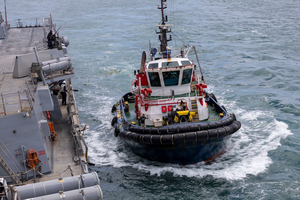 A harbor tugboat with red and white superstructure and black rubber fendering is operating alongside a gray naval warship. The tugboat appears to be assisting the larger vessel, likely during a docking or maneuvering operation. Several crew members are visible on both vessels, and the water shows active wake from the tugboat's propulsion.