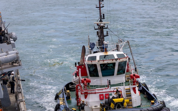 A harbor tugboat with red and white superstructure and black rubber fendering is operating alongside a gray naval warship. The tugboat appears to be assisting the larger vessel, likely during a docking or maneuvering operation. Several crew members are visible on both vessels, and the water shows active wake from the tugboat's propulsion.