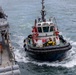 A harbor tugboat with red and white superstructure and black rubber fendering is operating alongside a gray naval warship. The tugboat appears to be assisting the larger vessel, likely during a docking or maneuvering operation. Several crew members are visible on both vessels, and the water shows active wake from the tugboat's propulsion.