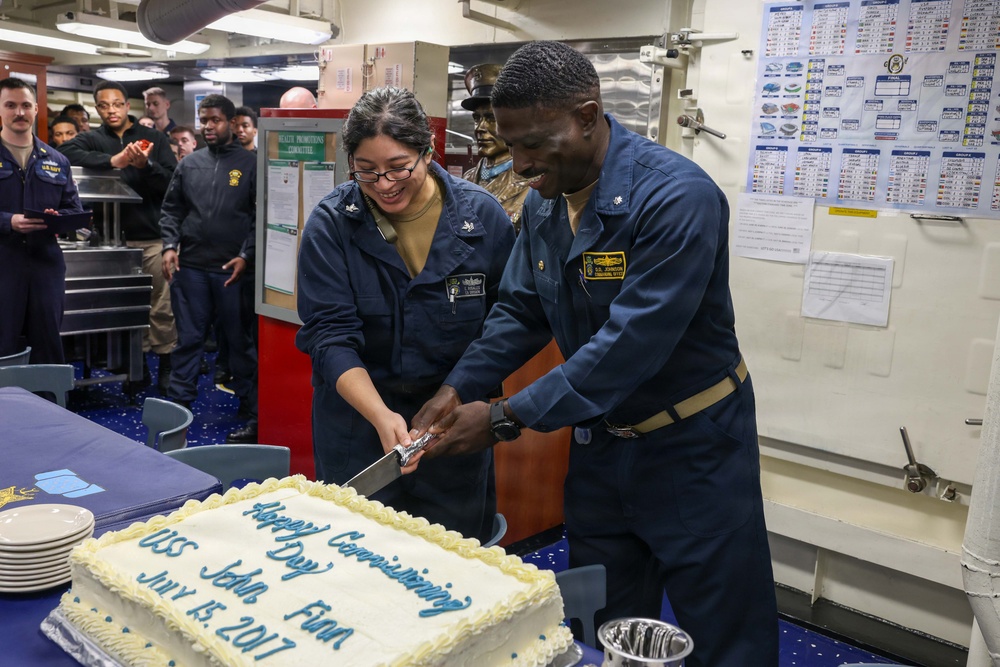 Two U.S. Navy sailors in blue coverall uniforms are cutting a white celebration cake aboard a naval vessel. The cake reads 'Happy Commissioning Day USS John Finn July 15, 2017,' marking the commissioning anniversary of the USS John Finn (DDG-113). A group of fellow sailors observes the ceremony in the background of what appears to be a mess deck or common area.