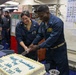 Two U.S. Navy sailors in blue coverall uniforms are cutting a white celebration cake aboard a naval vessel. The cake reads 'Happy Commissioning Day USS John Finn July 15, 2017,' marking the commissioning anniversary of the USS John Finn (DDG-113). A group of fellow sailors observes the ceremony in the background of what appears to be a mess deck or common area.
