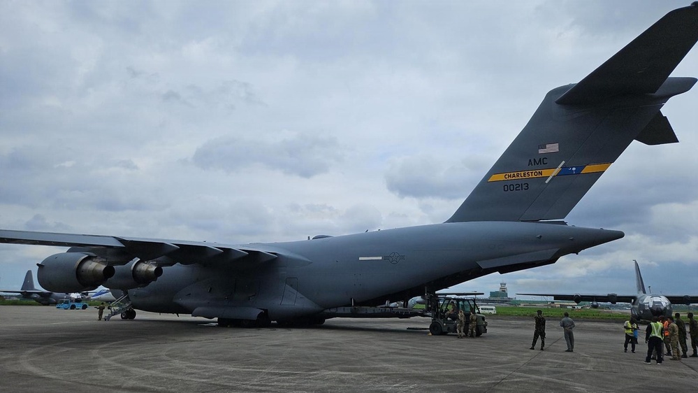 A U.S. Air Force C-17 Globemaster III cargo aircraft, tail number 00213, assigned to AMC Charleston, is parked on an airport tarmac. Ground crew personnel and support vehicles are visible near the rear of the aircraft, likely conducting loading or unloading operations. The setting appears to be an overseas or tropical airfield based on the vegetation and airport infrastructure visible in the background.