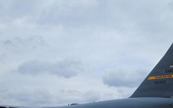 A U.S. Air Force C-17 Globemaster III cargo aircraft, tail number 00213, assigned to AMC Charleston, is parked on an airport tarmac. Ground crew personnel and support vehicles are visible near the rear of the aircraft, likely conducting loading or unloading operations. The setting appears to be an overseas or tropical airfield based on the vegetation and airport infrastructure visible in the background.