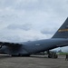 A U.S. Air Force C-17 Globemaster III cargo aircraft, tail number 00213, assigned to AMC Charleston, is parked on an airport tarmac. Ground crew personnel and support vehicles are visible near the rear of the aircraft, likely conducting loading or unloading operations. The setting appears to be an overseas or tropical airfield based on the vegetation and airport infrastructure visible in the background.