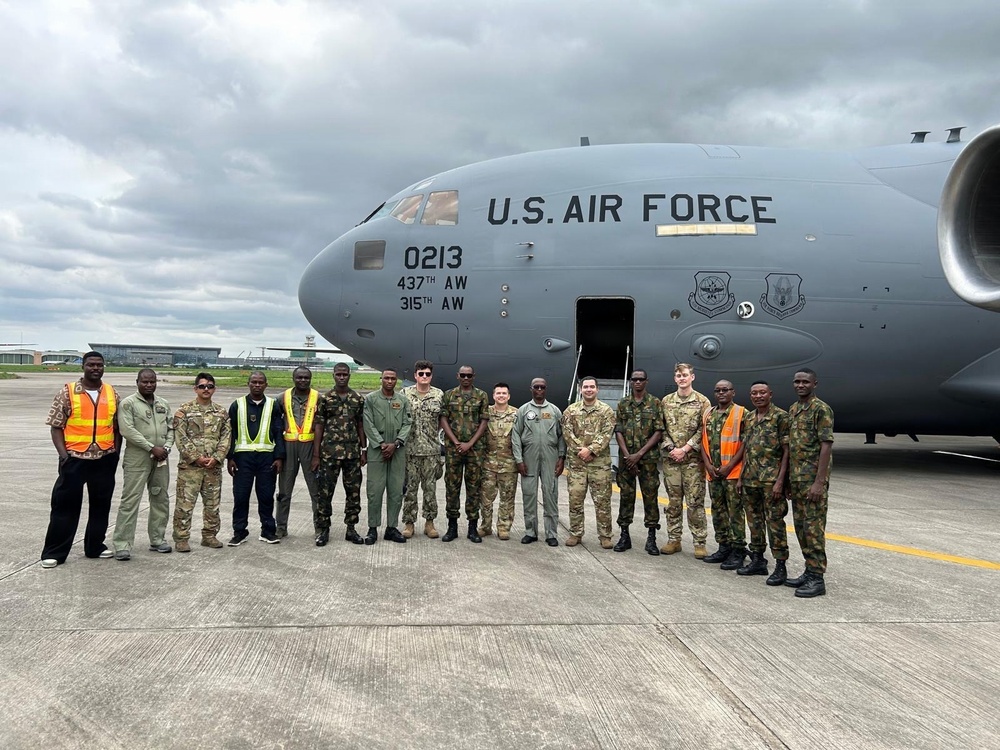 A group of approximately 18 military personnel and civilian workers pose in front of a U.S. Air Force C-17 Globemaster III aircraft on an airport tarmac. The aircraft bears tail number 0213 and is assigned to both the 437th Airlift Wing and 315th Airlift Wing. The group includes U.S. Air Force personnel in various uniforms alongside what appear to be host-nation military members in camouflage uniforms, suggesting a joint or partnership engagement.