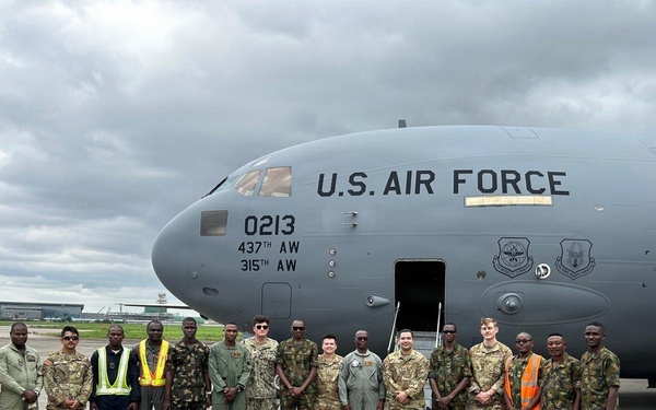 A group of approximately 18 military personnel and civilian workers pose in front of a U.S. Air Force C-17 Globemaster III aircraft on an airport tarmac. The aircraft bears tail number 0213 and is assigned to both the 437th Airlift Wing and 315th Airlift Wing. The group includes U.S. Air Force personnel in various uniforms alongside what appear to be host-nation military members in camouflage uniforms, suggesting a joint or partnership engagement.