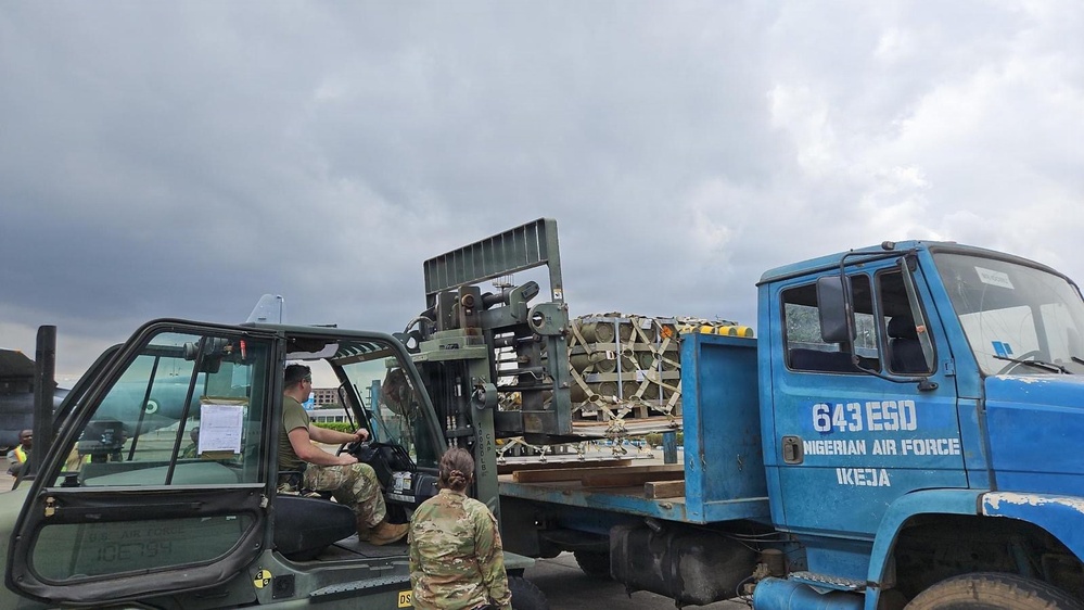 Military personnel are conducting a cargo loading or unloading operation on what appears to be an airfield. A military forklift is being used to transfer palletized cargo onto or from a blue flatbed truck marked '643 ESD Nigerian Air Force Ikeja'. The scene takes place under overcast skies with aircraft visible in the background.
