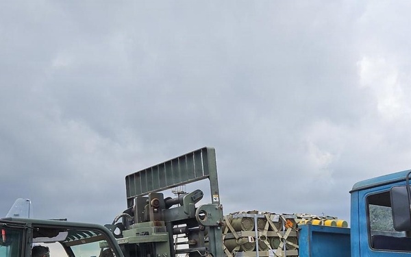 Military personnel are conducting a cargo loading or unloading operation on what appears to be an airfield. A military forklift is being used to transfer palletized cargo onto or from a blue flatbed truck marked '643 ESD Nigerian Air Force Ikeja'. The scene takes place under overcast skies with aircraft visible in the background.