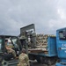 Military personnel are conducting a cargo loading or unloading operation on what appears to be an airfield. A military forklift is being used to transfer palletized cargo onto or from a blue flatbed truck marked '643 ESD Nigerian Air Force Ikeja'. The scene takes place under overcast skies with aircraft visible in the background.