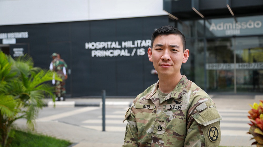 A U.S. Army soldier with the name tape 'KOO' and rank of Staff Sergeant stands in front of a building identified as 'Hospital Militar Principal' with a partial text reading 'IS'. The soldier is wearing OCP (Operational Camouflage Pattern) uniform with a U.S. Army tape, a unit patch on the left sleeve (appears to be 2nd Infantry Division), and a rank insignia. In the background, another military figure in a different camouflage uniform is visible near the building entrance.
