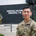 A U.S. Army soldier with the name tape 'KOO' and rank of Staff Sergeant stands in front of a building identified as 'Hospital Militar Principal' with a partial text reading 'IS'. The soldier is wearing OCP (Operational Camouflage Pattern) uniform with a U.S. Army tape, a unit patch on the left sleeve (appears to be 2nd Infantry Division), and a rank insignia. In the background, another military figure in a different camouflage uniform is visible near the building entrance.