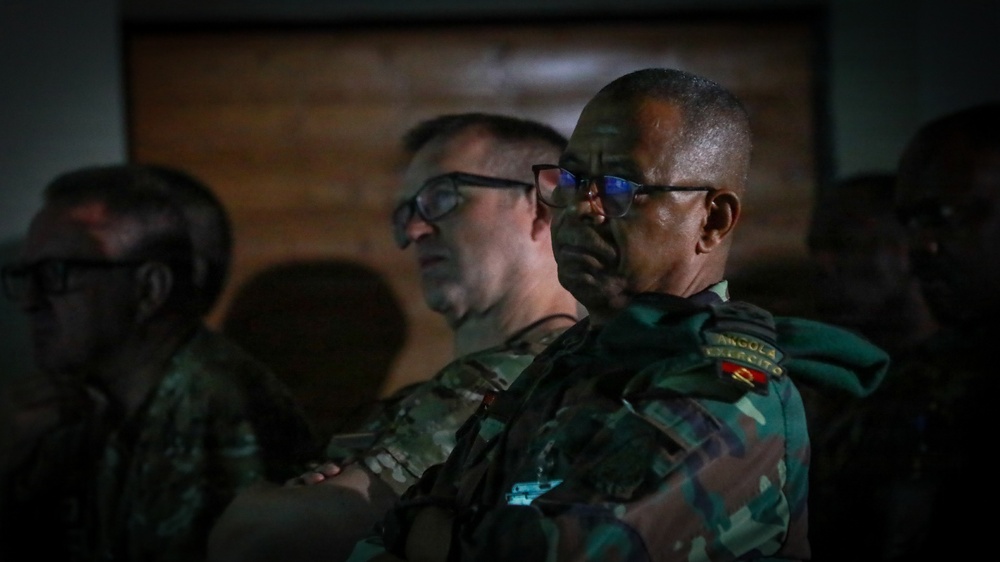 The image shows military personnel in camouflage uniforms attending what appears to be a briefing or presentation in a dimly lit room. At least two prominent figures are visible in the foreground, one wearing a patch that reads 'Angola Exército' (Angola Army), suggesting a multinational military gathering or joint exercise. The low lighting and focused expressions suggest a formal military setting.