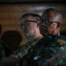 The image shows military personnel in camouflage uniforms attending what appears to be a briefing or presentation in a dimly lit room. At least two prominent figures are visible in the foreground, one wearing a patch that reads 'Angola Exército' (Angola Army), suggesting a multinational military gathering or joint exercise. The low lighting and focused expressions suggest a formal military setting.