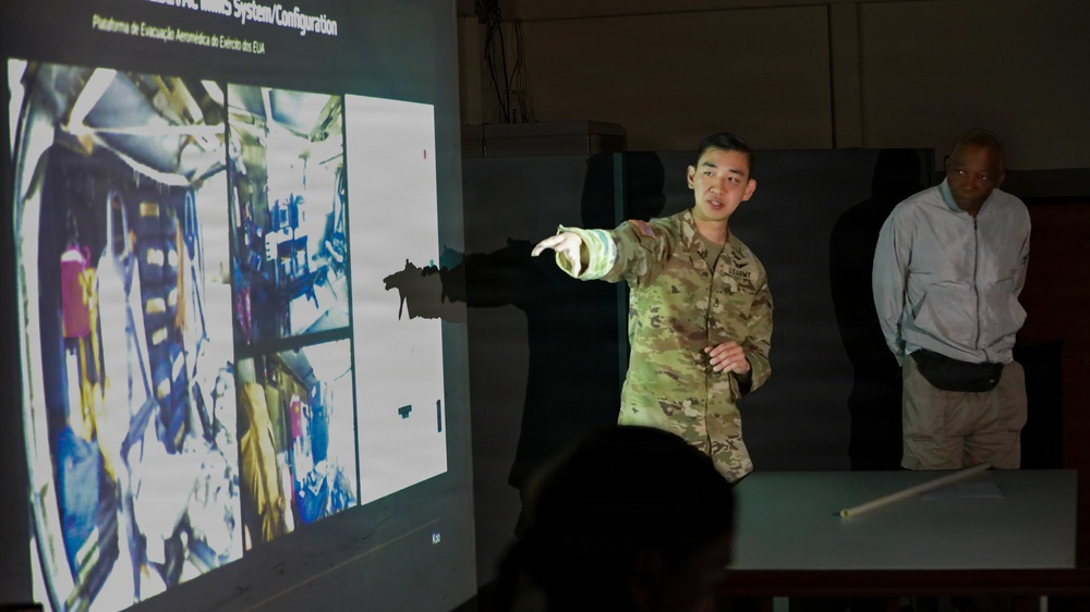 A soldier in US Army camouflage uniform is presenting a briefing, pointing at a projected slide showing equipment images. The slide title references 'MEDEVAC MIMS System/Configuration' with Portuguese subtitle 'Plataforma de Evacuação Aeromédica do Exército dos EUA' (US Army Aeromedical Evacuation Platform). A second individual in civilian clothing stands in the background observing the presentation.