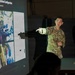 A soldier in US Army camouflage uniform is presenting a briefing, pointing at a projected slide showing equipment images. The slide title references 'MEDEVAC MIMS System/Configuration' with Portuguese subtitle 'Plataforma de Evacuação Aeromédica do Exército dos EUA' (US Army Aeromedical Evacuation Platform). A second individual in civilian clothing stands in the background observing the presentation.
