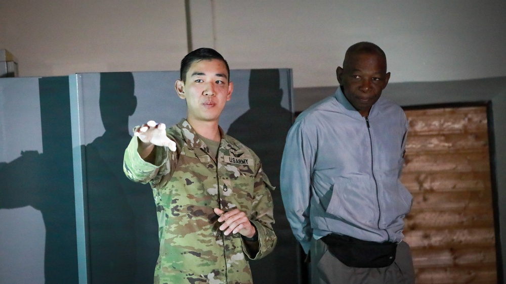 Two individuals are standing indoors in what appears to be a dimly lit facility or building. One person is wearing a U.S. Army camouflage uniform and appears to be gesturing or pointing, while the second person is dressed in civilian athletic wear. Their shadows are cast prominently on the wall behind them.
