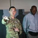 Two individuals are standing indoors in what appears to be a dimly lit facility or building. One person is wearing a U.S. Army camouflage uniform and appears to be gesturing or pointing, while the second person is dressed in civilian athletic wear. Their shadows are cast prominently on the wall behind them.