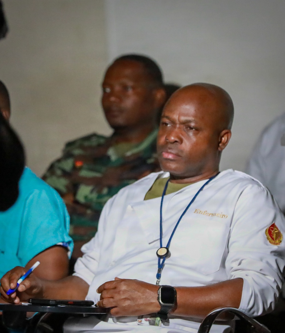 The image shows several individuals seated in what appears to be a meeting or briefing setting. The foreground subject is a bald man wearing a white medical coat labeled 'Enfermeiro' (Portuguese for 'Nurse') with a military medical insignia patch on the sleeve and a blue lanyard. Behind him, a person in military camouflage uniform is visible, suggesting a mixed civilian-military medical context.