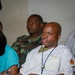 The image shows several individuals seated in what appears to be a meeting or briefing setting. The foreground subject is a bald man wearing a white medical coat labeled 'Enfermeiro' (Portuguese for 'Nurse') with a military medical insignia patch on the sleeve and a blue lanyard. Behind him, a person in military camouflage uniform is visible, suggesting a mixed civilian-military medical context.