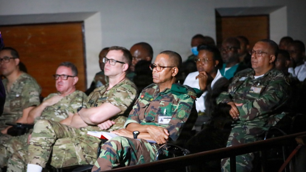 A group of military personnel in various camouflage uniforms are seated in what appears to be an auditorium or briefing room, attending a presentation or meeting. The audience includes individuals wearing different camouflage patterns, suggesting a multinational or joint military gathering. Civilians are also visible in the background rows.