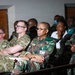 A group of military personnel in various camouflage uniforms are seated in what appears to be an auditorium or briefing room, attending a presentation or meeting. The audience includes individuals wearing different camouflage patterns, suggesting a multinational or joint military gathering. Civilians are also visible in the background rows.