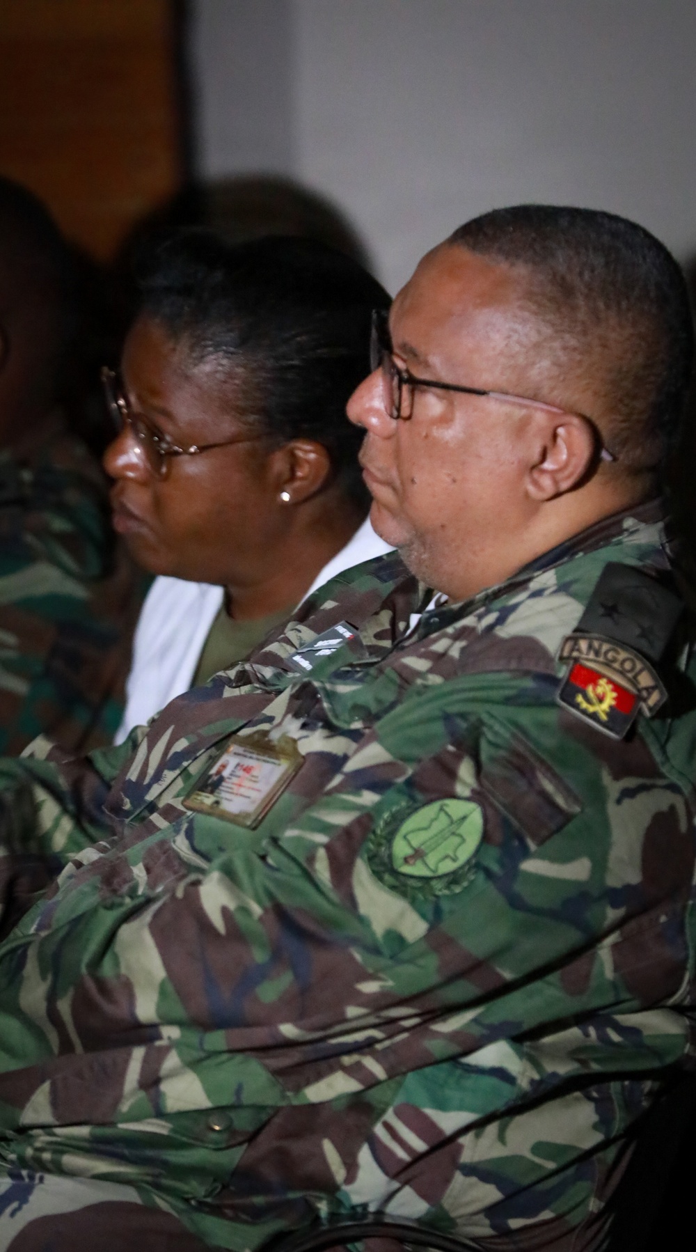 Two military personnel wearing Angolan Armed Forces camouflage uniforms are seated, appearing to attend a briefing or formal event. The foreground individual is a male wearing glasses with visible 'ANGOLA' shoulder patch, a unit patch on the sleeve, and an identification badge on the chest. A female soldier wearing glasses is visible in the background.