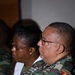 Two military personnel wearing Angolan Armed Forces camouflage uniforms are seated, appearing to attend a briefing or formal event. The foreground individual is a male wearing glasses with visible 'ANGOLA' shoulder patch, a unit patch on the sleeve, and an identification badge on the chest. A female soldier wearing glasses is visible in the background.