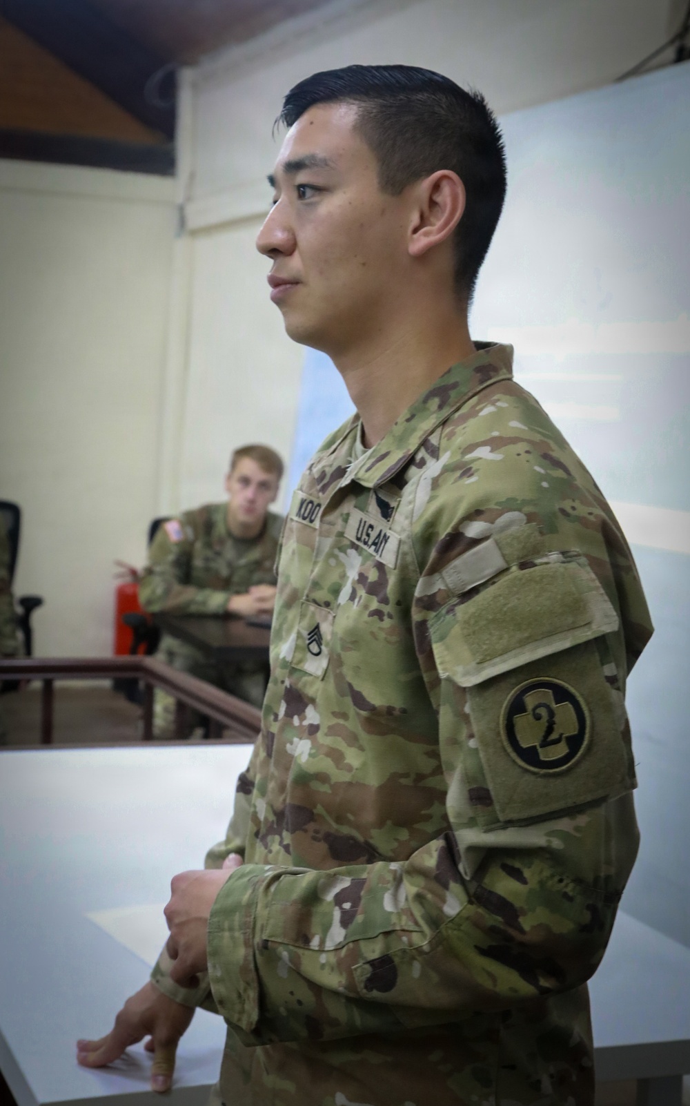 A U.S. Army soldier in Operational Camouflage Pattern (OCP) uniform is standing in what appears to be a classroom or briefing room. The soldier's name tape reads 'KOO' and displays a Staff Sergeant rank insignia along with a '2nd Infantry Division' shoulder sleeve insignia. A second soldier is visible seated in the background.